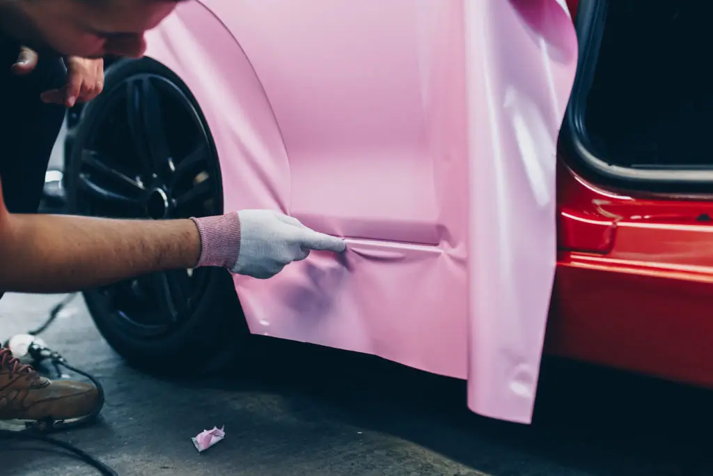 A close-up of a professional applying a pink vinyl wrap to the side panel of a red car, showcasing precision and attention to detail.