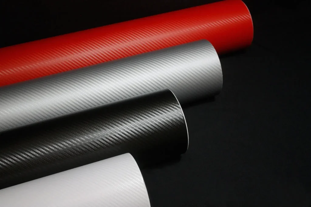 Four rolls of carbon fiber vinyl film in different colors (red, silver, black, and white) placed on a black background