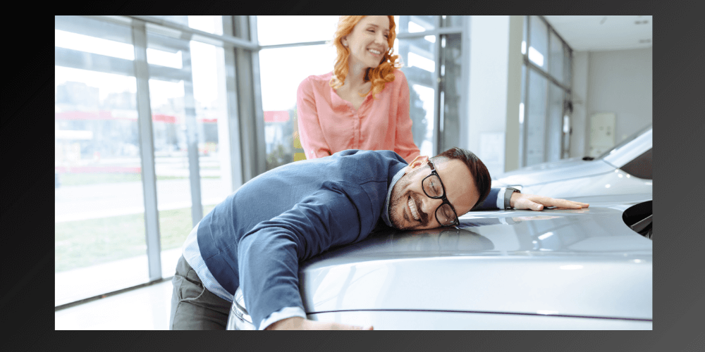  happy man in glasses hugging the hood of a car because he finds a new cool color a silver car with a smiling woman in the background.