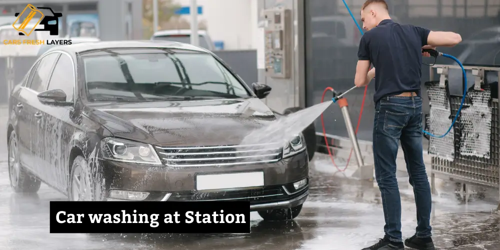 Person washing a dark sedan at a self-service car wash station using a high-pressure water hose