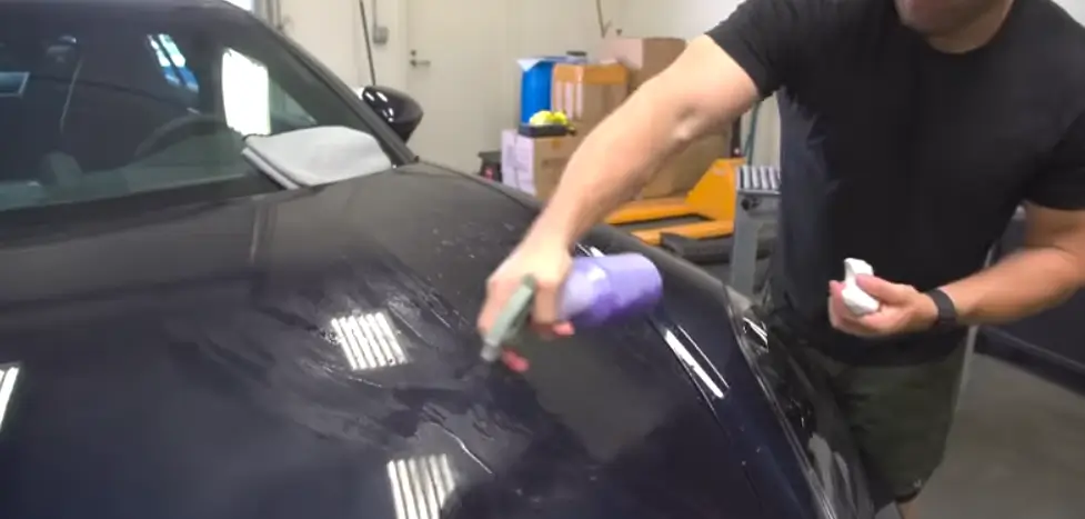 Person polishing or cleaning a car with a spray bottle, using a cloth and car detailing products in a garage.