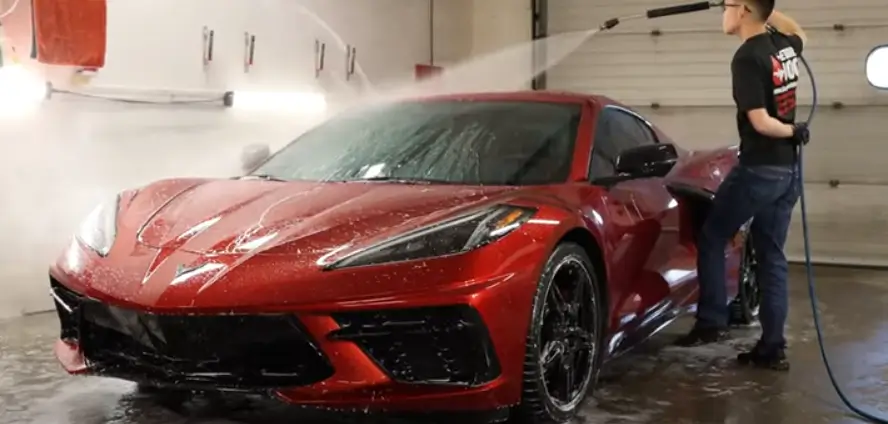 A red sports car being cleaned with a high-pressure water hose by a person in a car wash bay.