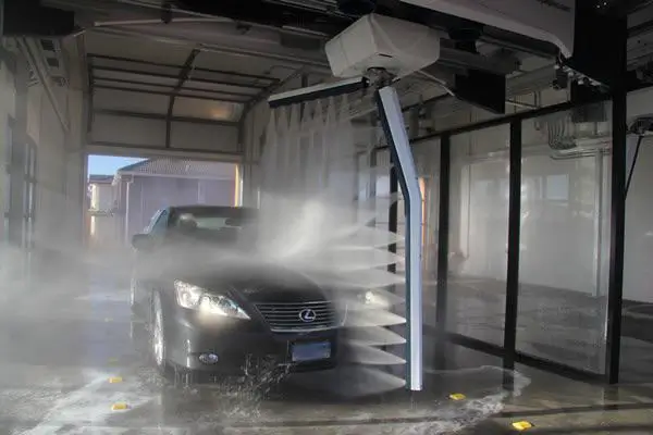 A black Lexus car in an automated car wash types with high-pressure water jets spraying from a rotating arm.