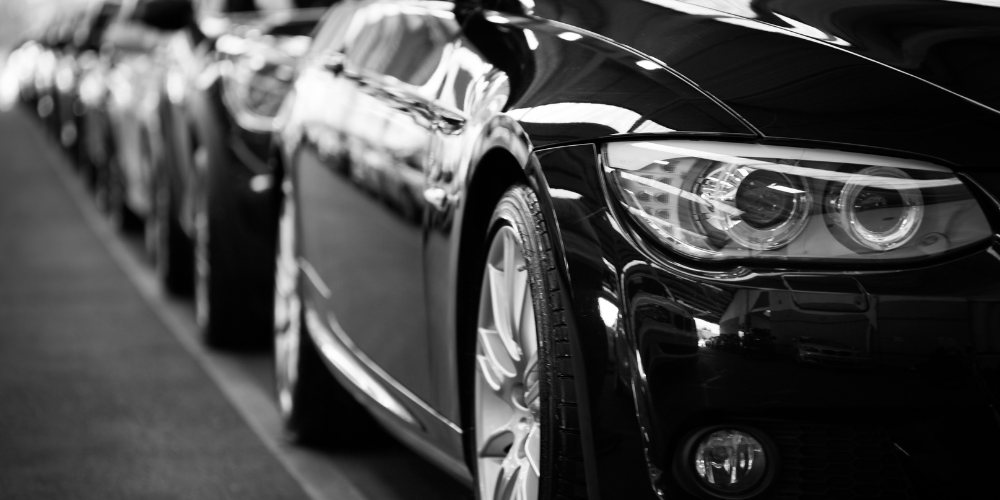 Close-up black-and-white photo of a sleek car with prominent headlights, part of a row of cars parked in a line