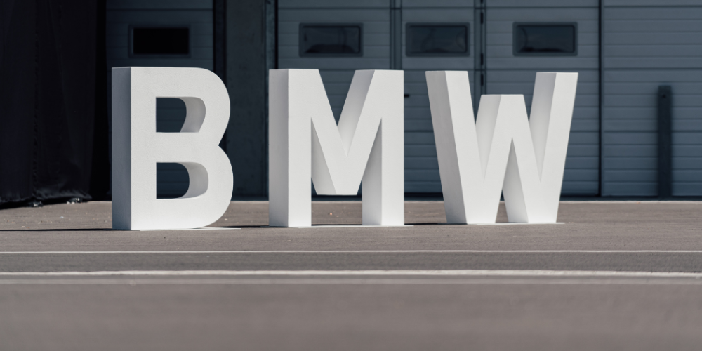 Large white 3D letters spelling 'BMW' placed outdoors on a paved surface with a building featuring garage doors in the background